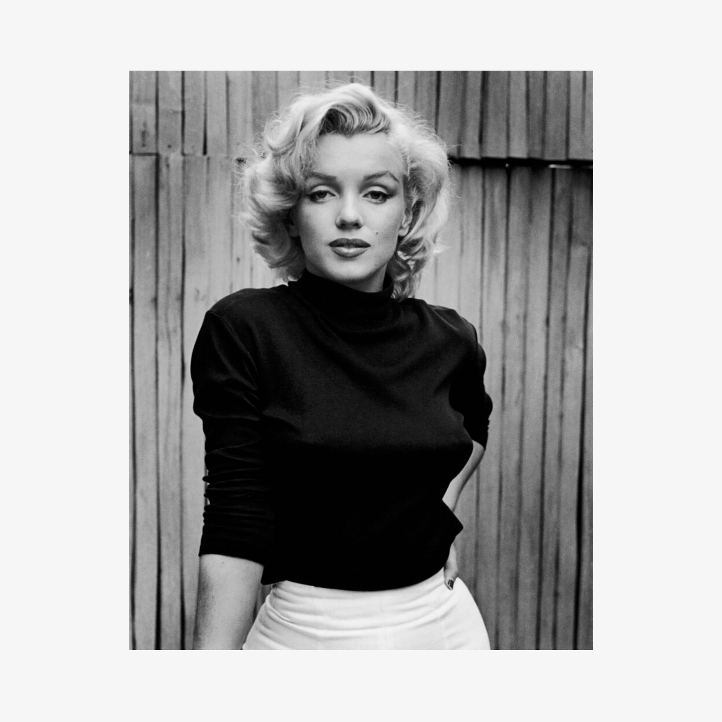 Black and white photo of a person wearing a black turtleneck and white skirt against a wooden fence.