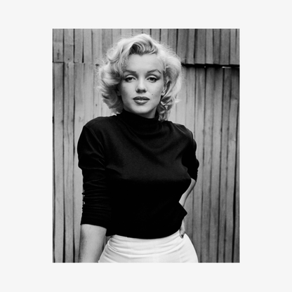 Black and white photo of a person wearing a black turtleneck and white skirt against a wooden fence.