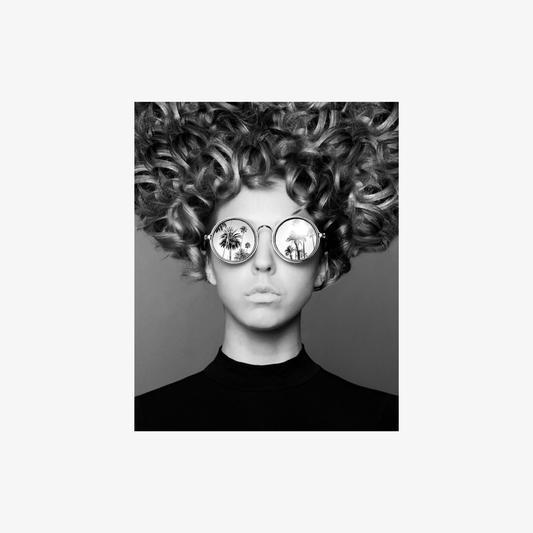 Person with curly hair wearing round sunglasses against a plain background