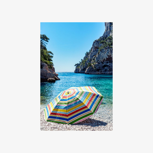 Colorful striped beach umbrella on a pebbly beach with turquoise water and rocky cliffs in the background.