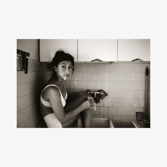 Black and white photo of a woman sitting on a toilet in a tiled bathroom.