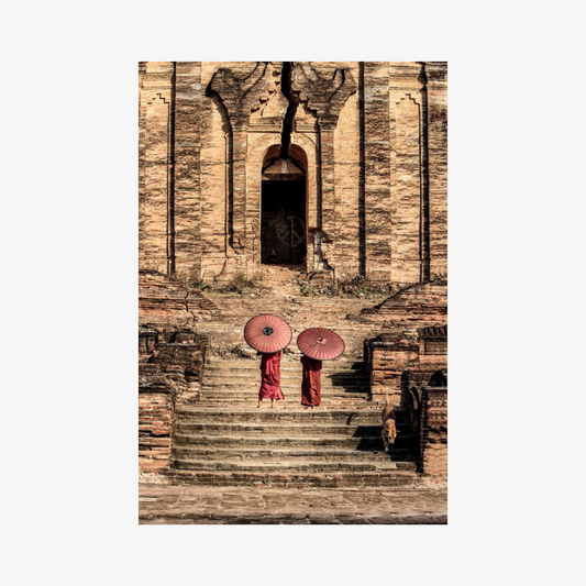 Two people in red robes holding pink umbrellas on stone steps in front of a temple.