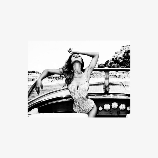 Black and white photo of a woman sitting on a boat with arms raised, nature in the background